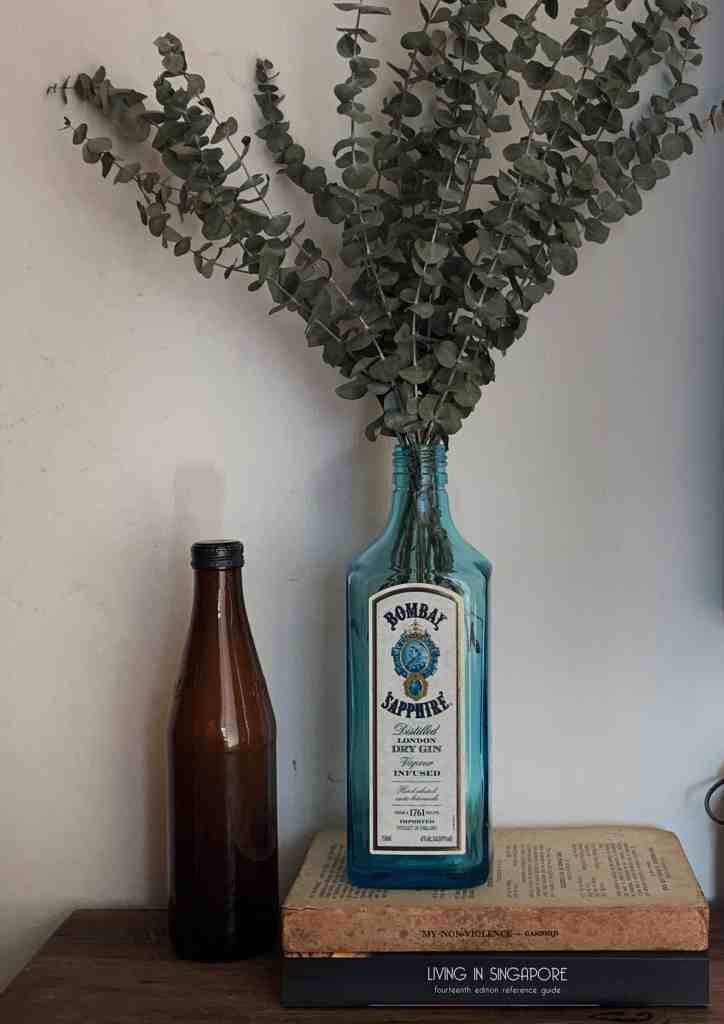 ユーカリの枝を飾る花瓶として再利用された空のボンベイ・サファイア・ジンのボトルが、茶色のガラス瓶と積み重ねられた本の横に置かれている。