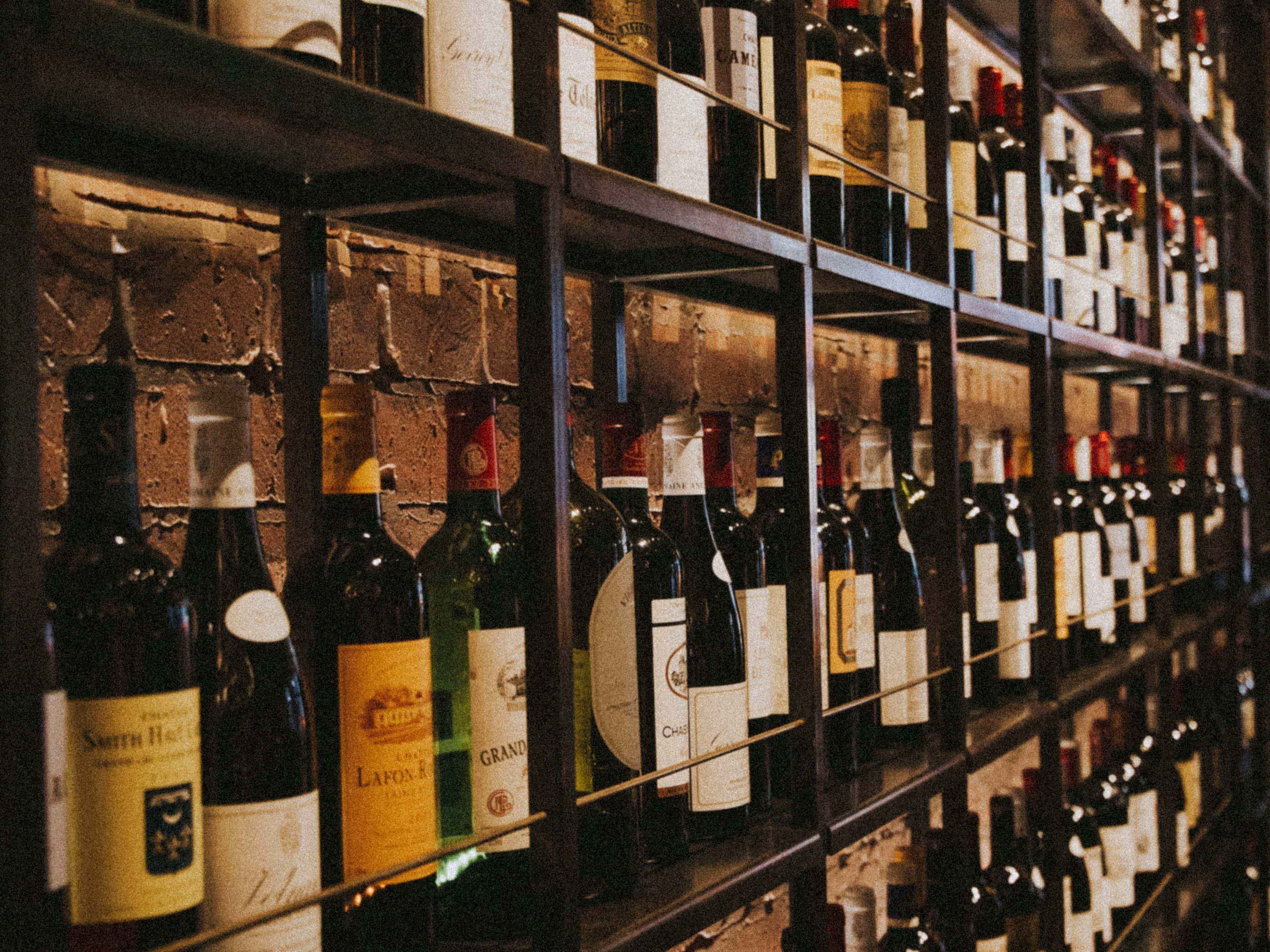Una toma ligeramente inclinada de un gran botellero repleto de botellas de vino tinto y blanco. Al fondo, una pared de ladrillos crea un ambiente clásico, similar al de una bodega.