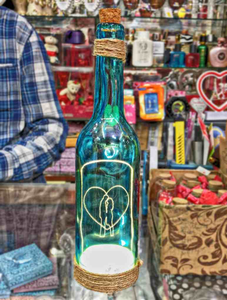 紐で巻かれ、内側から照らされた青いガラス瓶には、ぼやけた小売店の棚の背景に、2 人の人物が描かれた光るハートの輪郭が映し出されています。