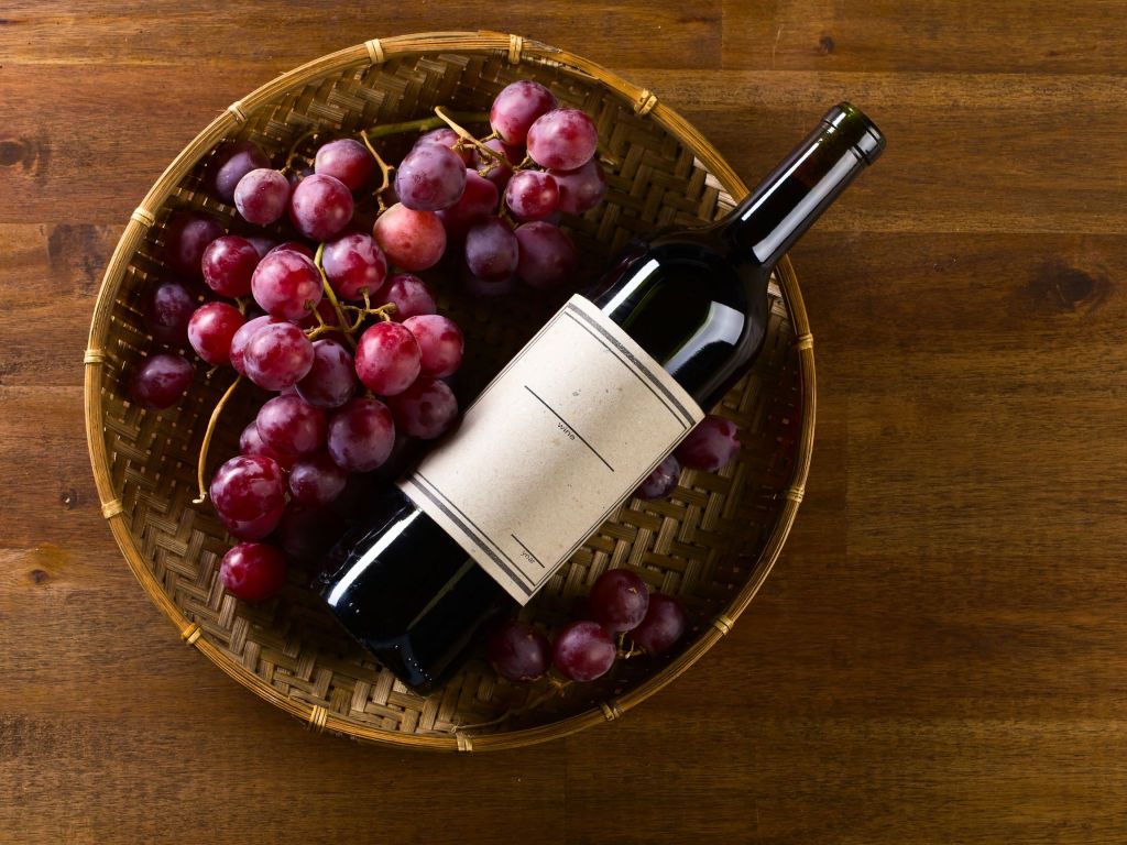 A bottle of red wine lying across a woven bamboo basket filled with fresh red and purple grapes on a wooden table.