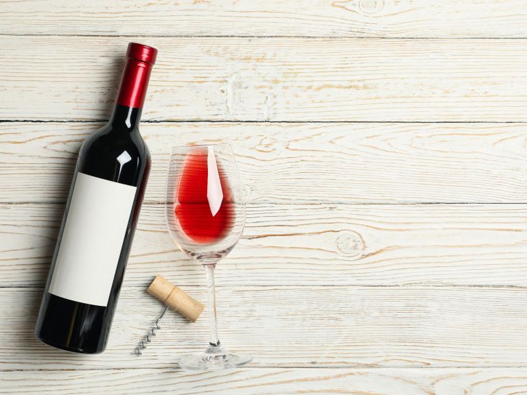 A single bottle of red wine with a blank label, a half-filled glass of red wine, and a corkscrew on a light wooden table.