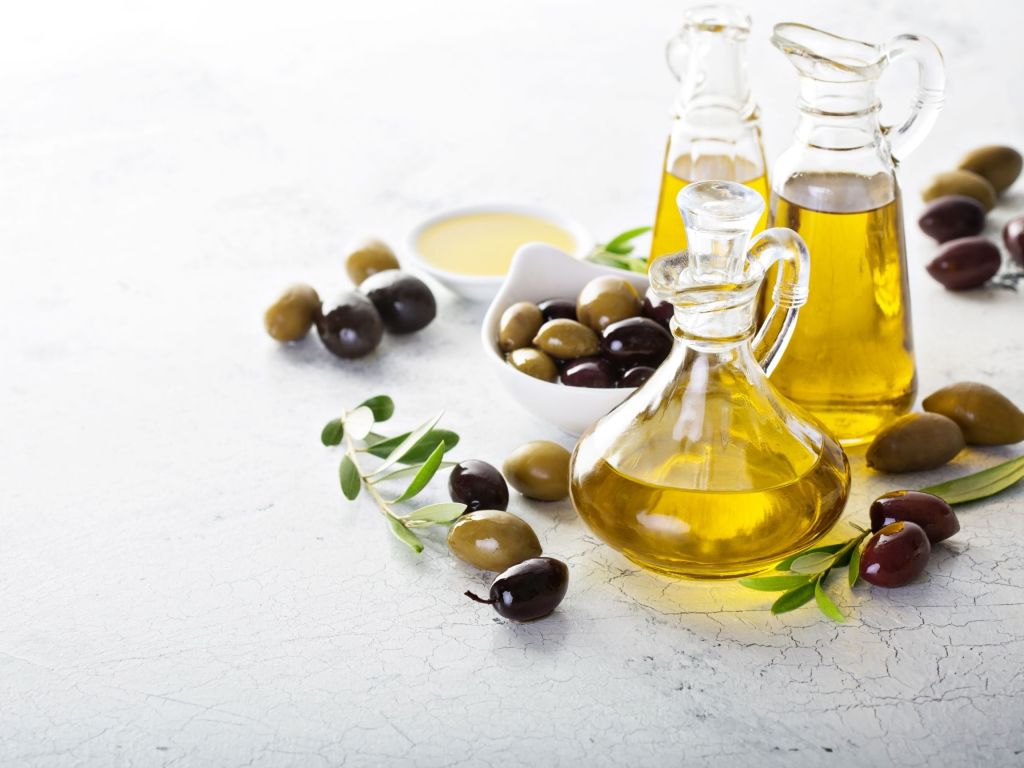 Various bottles and dispensers of olive oil in shades of golden yellow, accompanied by green and black olives on a light background.