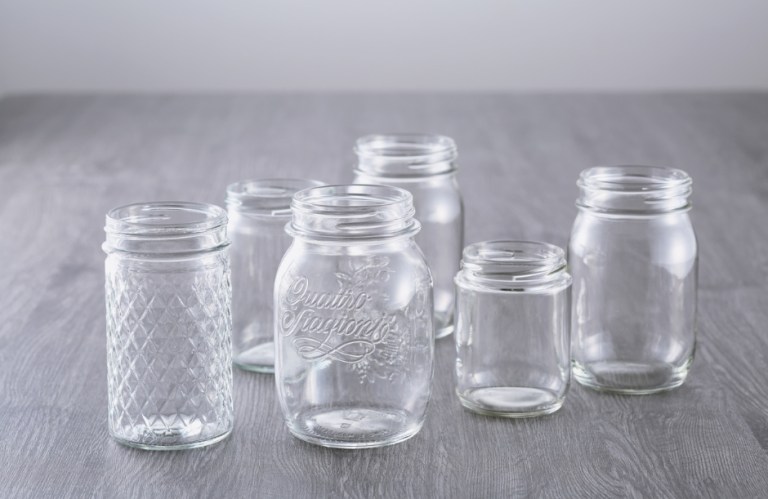 Five empty clear glass jars in different designs, including textured, embossed "Quattro Stagioni," and plain styles, arranged on a wooden table.