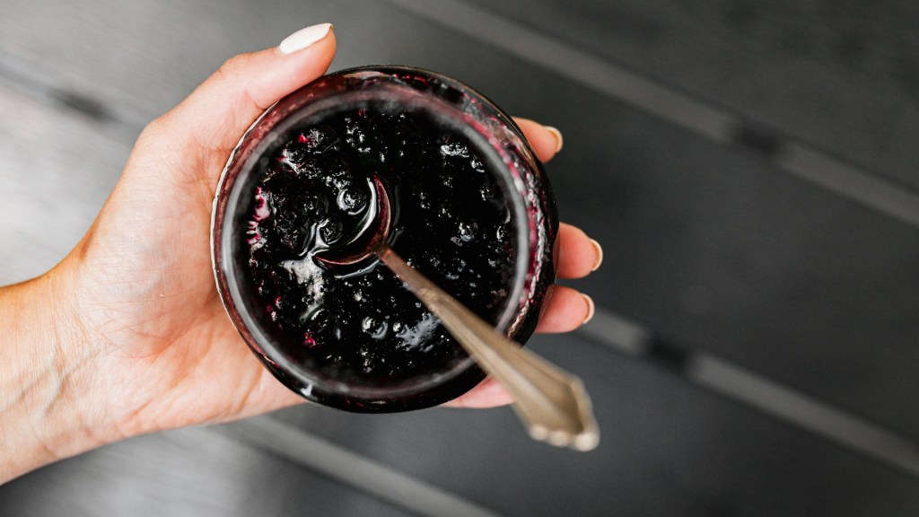 Close-up of a hand holding an open jar of dark purple-black berry jam with a silver spoon dipped in, some jam smeared on the rim.
