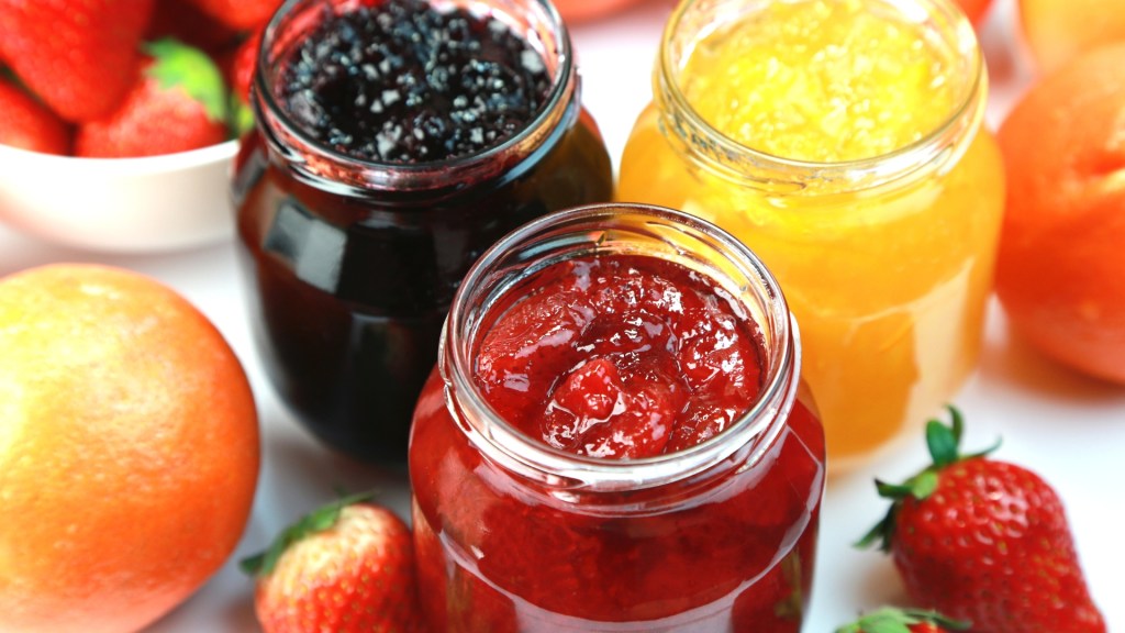Four open glass jars showing chunky strawberry jam, dark blackberry or blueberry jam, and citrus marmalade, surrounded by fresh strawberries and oranges.