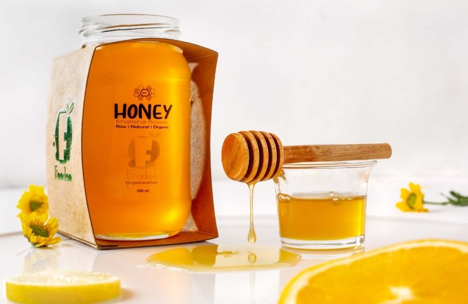 A jar of bright golden honey in eco-friendly cardboard packaging labeled “HONEY – Raw | Natural | Organic” (500 ml), accompanied by a wooden honey dipper dripping honey into a small glass bowl, with lemon slices and yellow flowers on a white surface.