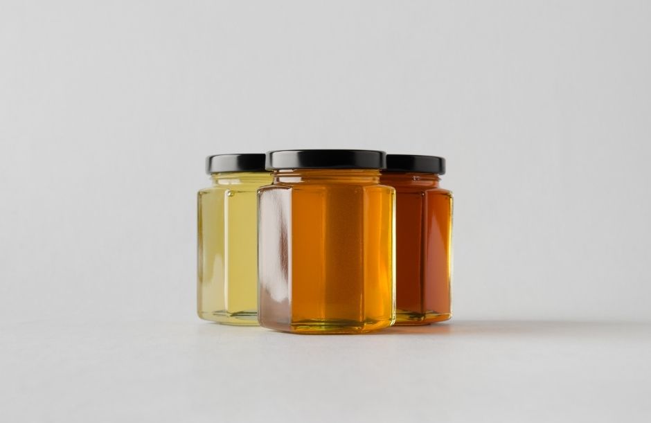 Three hexagonal glass jars with black lids placed side by side on a clean white background, containing honey in a gradient from very light golden (almost clear) on the left, medium amber in the middle, to deep reddish-brown on the right.