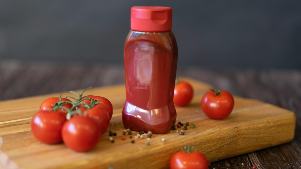 Red plastic ketchup bottle with flip-top lid on a wooden board, surrounded by fresh cherry tomatoes and peppercorns.