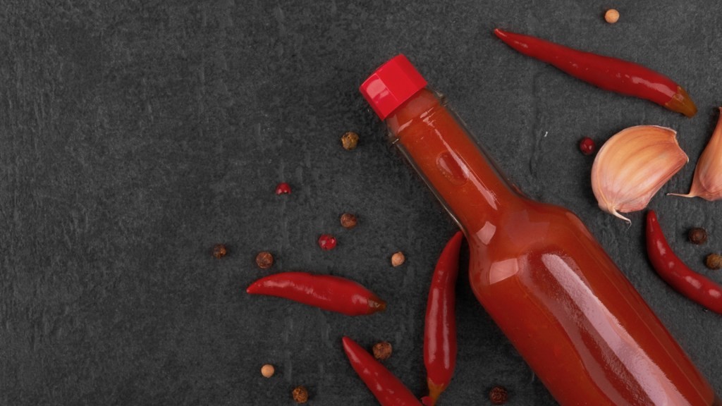 Bottle of red hot sauce with red cap lying on dark slate, surrounded by red chilies, garlic, and spices.