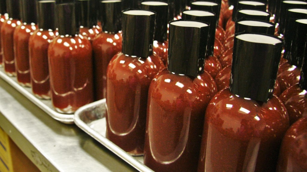 Many identical bottles filled with red hot sauce, capped in black, arranged in rows on metal trays in a factory setting.