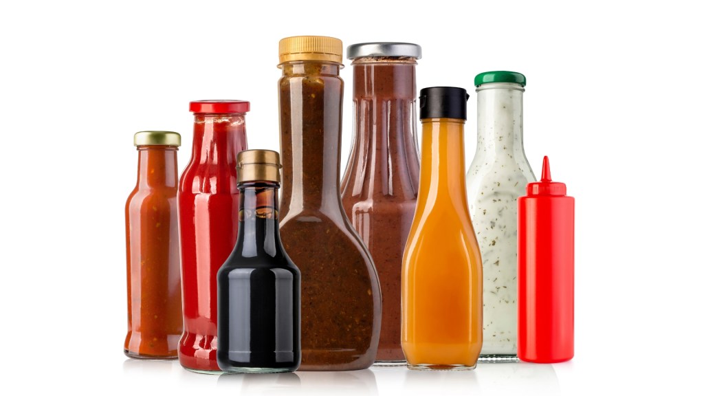 Nine assorted condiment bottles in different shapes and colors (red, brown, orange, white, dark) lined up on a white background.