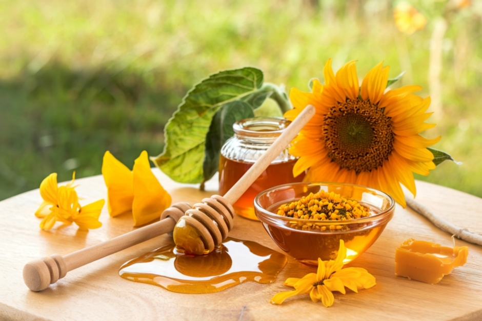 Rustic wooden table with a jar of honey, wooden honey dipper dripping honey, fresh sunflowers, a bowl of bright yellow bee pollen, and a piece of raw beeswax under warm natural light.
