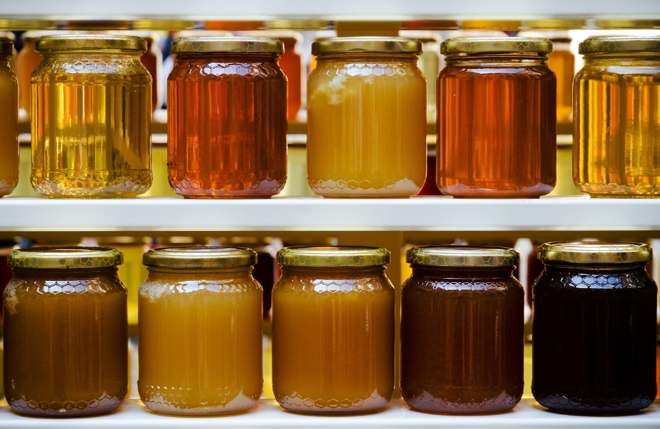 Two shelves displaying a gradient of honey jars in glass hexagon containers, ranging from pale golden and almost transparent to deep amber, reddish-brown, and nearly black honey.