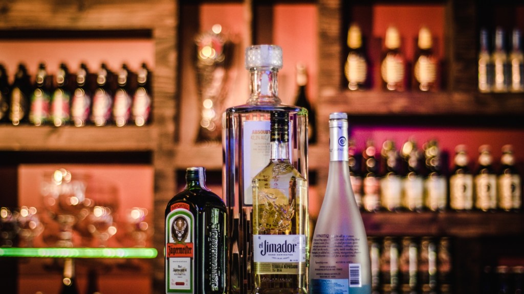 Four liquor bottles—Jägermeister, Absolut Vodka, El Jimador Tequila, and Hypnotiq—displayed on a glowing bar shelf with blurred colorful lights behind.
