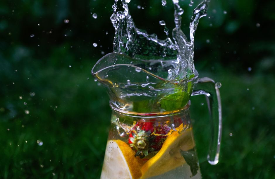 A glass pitcher of detox water infused with strawberries, lemon slices, apple, and fresh mint leaves, with water splashing over the rim against a green outdoor background