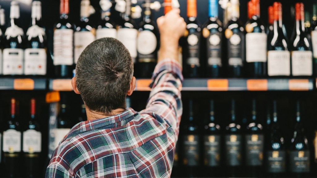 Back view of a man in a plaid shirt reaching for a bottle on a high shelf in a liquor store aisle filled with wine and spirits.