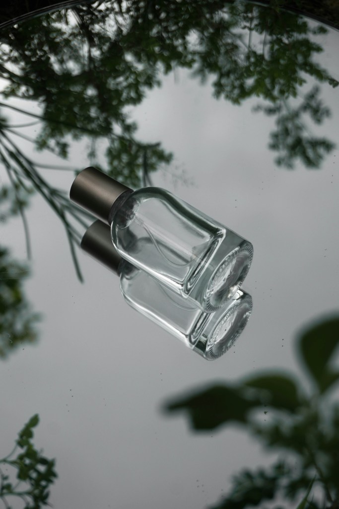 An empty clear glass mist spray bottle with a metallic cap resting on a mirror with tree reflections