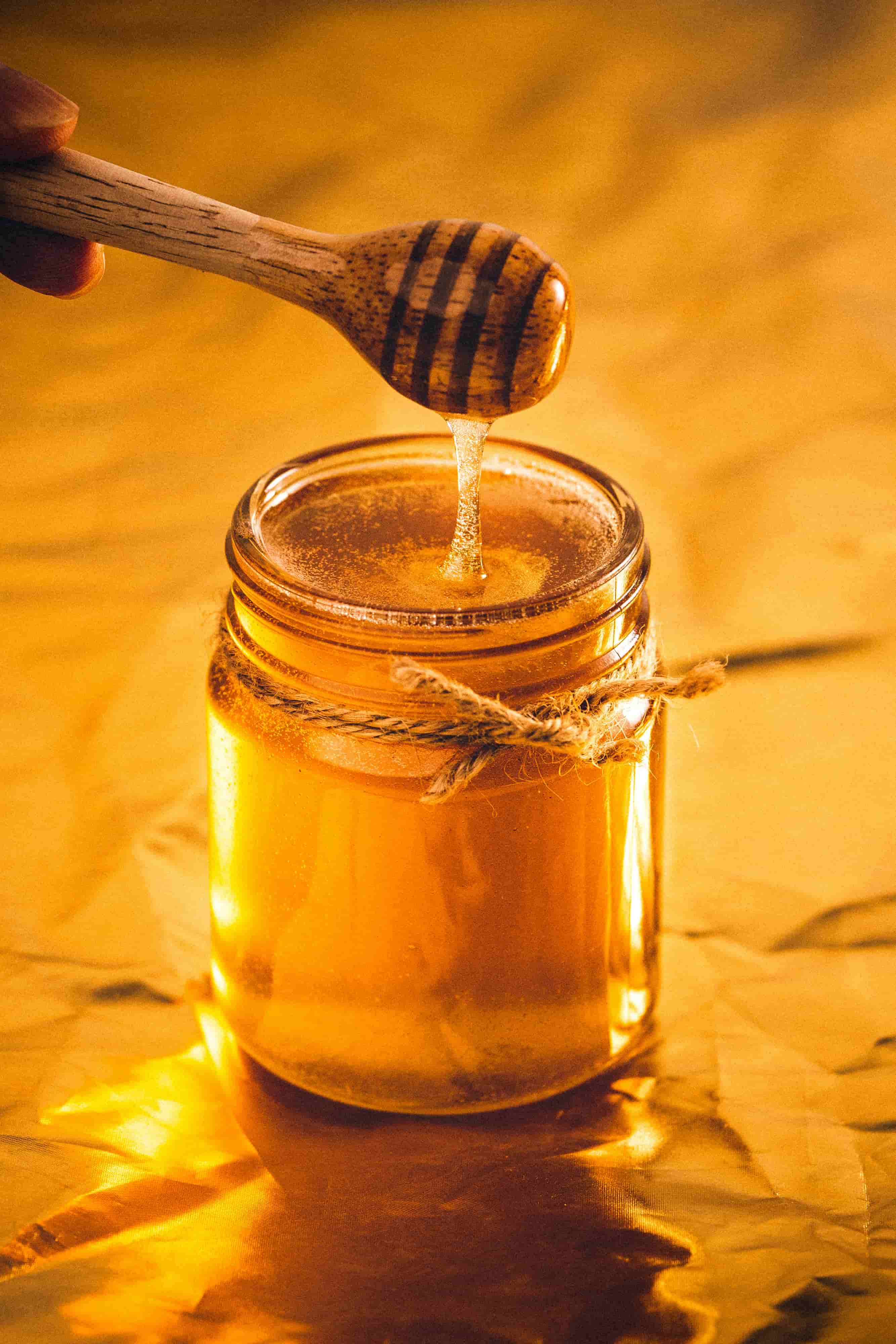 Golden honey dripping from wooden honey dipper into glass jar with warm lighting