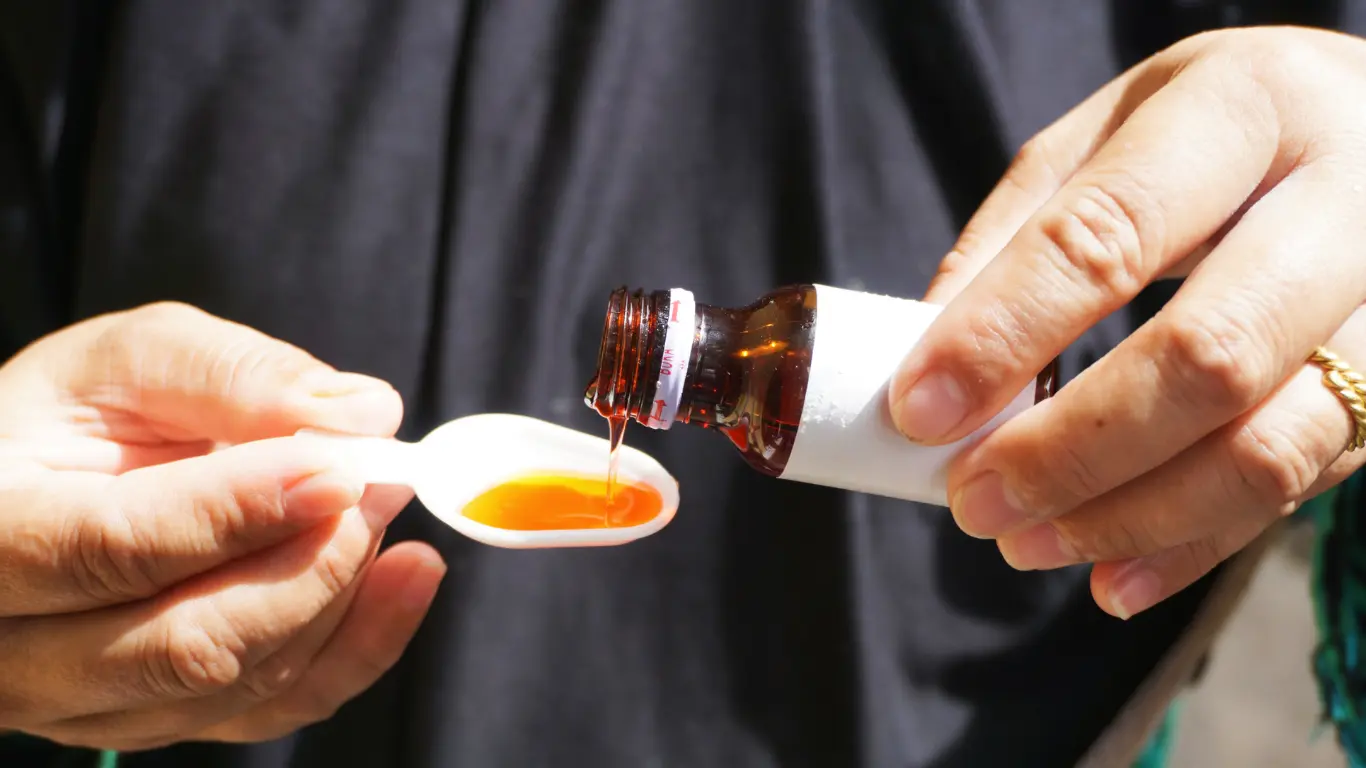 Hands pouring orange liquid syrup from a small amber bottle with a white tamper-evident seal into a plastic spoon