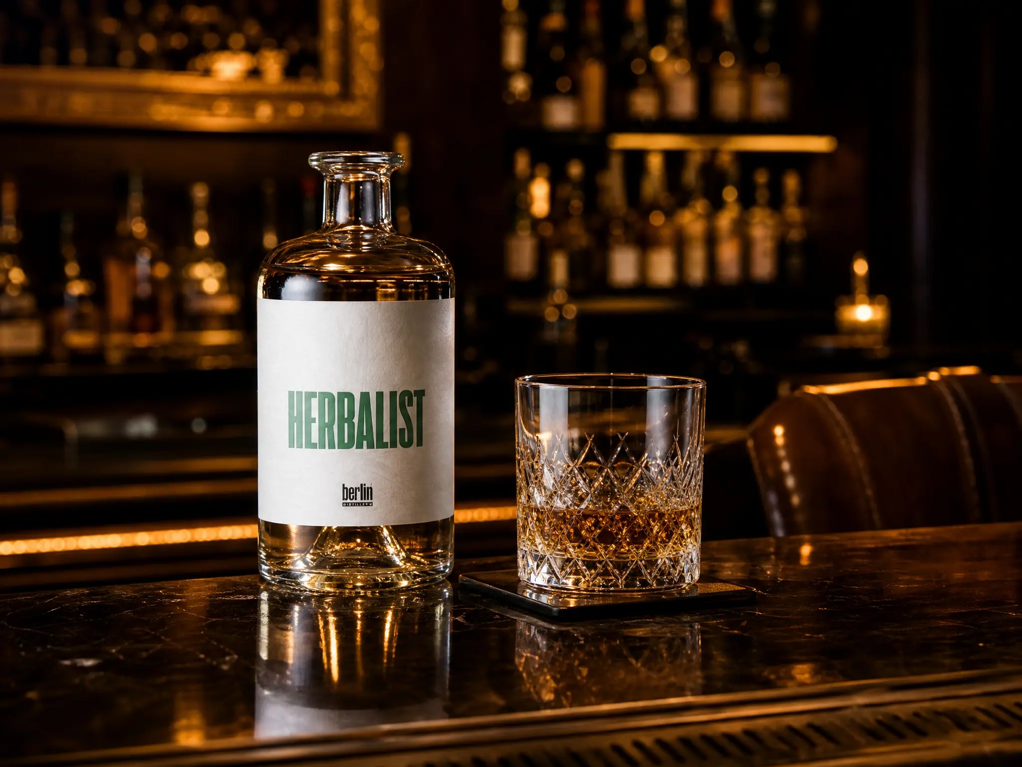 Clear empty Herbalist liquor glass bottle next to diamond cut crystal rocks glass with amber spirit on wooden bar counter, warm back bar bokeh lighting, Berlin Packaging APAC bulk wholesale herbal liqueur bottle